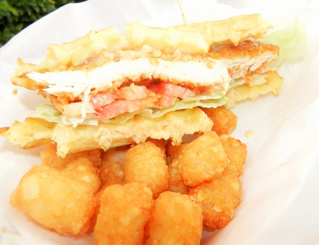 Mouthwatering Crunchy golden Chicken and Waffle Sandwich from Cletus' Chicken Shack with glaze maple syrup mayo on a bed of bright green lettuce and red tomatoes.