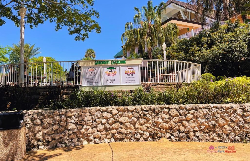 Busch Gardens Tampa Giraffe Bar Treetop Kitchen and Oasis Pizza with signage for Serengeti Overlook on the fence surrounded by greenery and rock wall. Keep reading to learn more about what to eat at Busch Gardens Tampa.