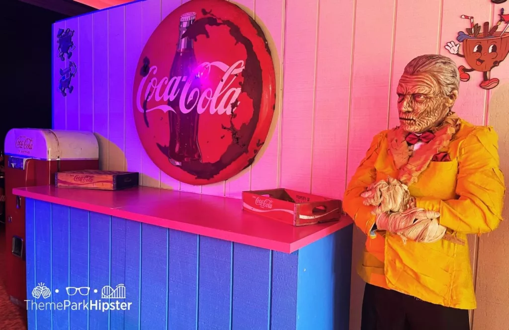 Retro style dead zombie like waiter standing beside a vintage looking serving area at Dead Coconut Club CityWalk during 2023 Halloween Horror Nights HHN 32 at Universal Studios Orlando. Keep reading to learn more about Halloween at Universal.