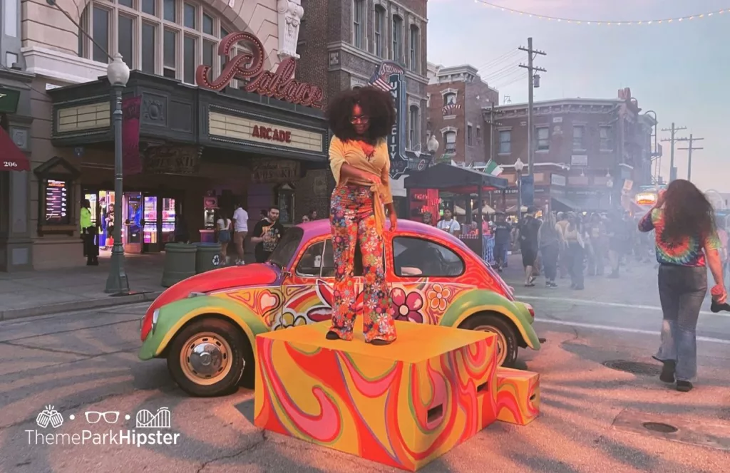 Vamp 69 Scare Zone with woman on top of block beside a vintage car in hippie colorful clothing with flowers during 2023 Halloween Horror Nights HHN 32 at Universal Studios Orlando. Keep reading to discover more about Halloween at Universal. 